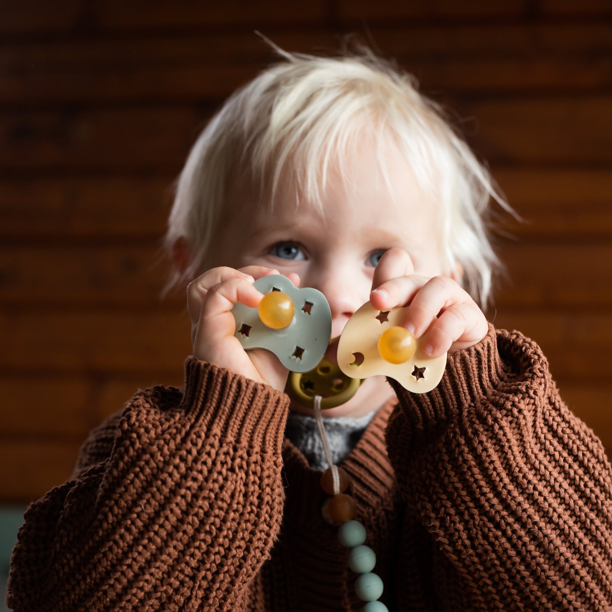Child holding a pacifier and teething ring in front of their face, wearing a brown knit cardigan.