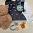 Baby items including a teddy bear, pacifiers, and a teething ring on a neutral background