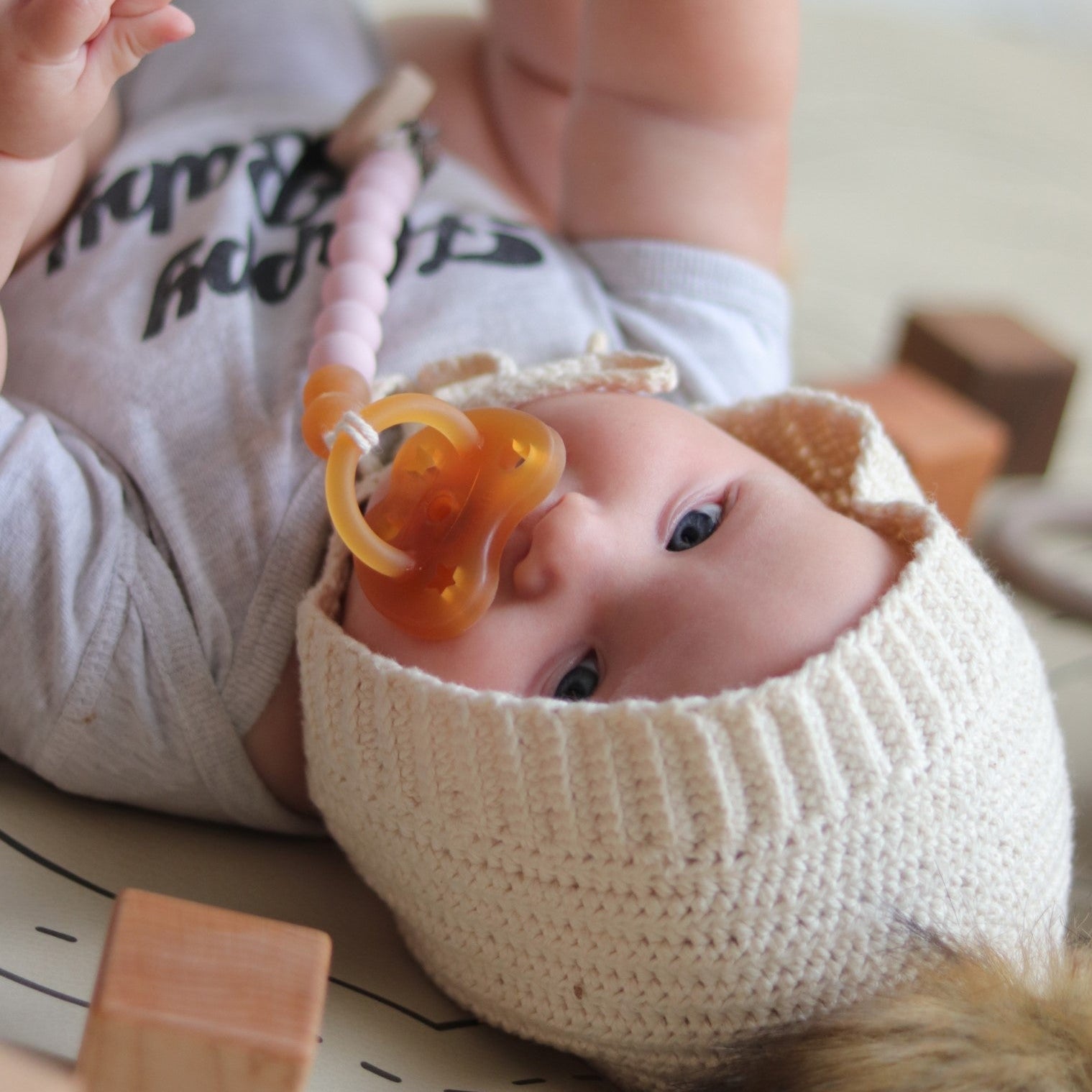 Baby wearing a white knit hat and holding a pacifier, with wooden blocks in the background.